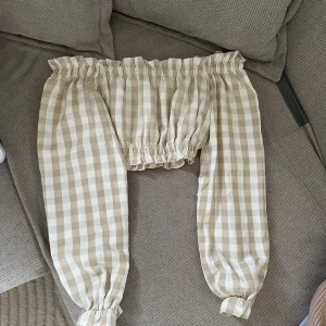 Beige och vit rutig offshoulder blus - Charmig offshoulder blus i beige och vitt rutigt mönster. Blusen har långa ärmar med elastiska muddar och en elastisk kant upptill för en bekväm passform. Perfekt för en avslappnad och stilren look.