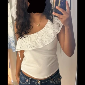 Vit oneshoulder topp från Hollister - Säljer en söt vit oneshoulder topp från Hollister med en bred volangdetalj. Toppen är ärmlös och har ett broderat mönster på volangen. Perfekt för sommaren!