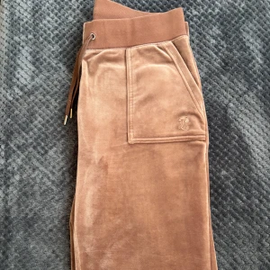 Ljus bruna Juicy Couture mjukisbyxor  - Snygga bruna mjukisbyxor i mjukt material med ribbad midja och snörning. Byxorna har praktiska fickor fram och bak. Perfekta för en avslappnad stil.