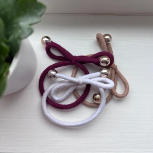 Hårsnoddar med rosett och metalldetaljer - Säljer tre hårsnoddar i olika färger: vinröd, vit och beige. Varje hårband har en söt rosett och är dekorerat med två metallkulor. Perfekt för att ge en extra touch till din frisyr.