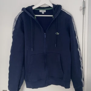 Mörkblå hoodie från Lacoste - Säljer en mörkblå hoodie från Lacoste med dragkedja och huva. Tröjan har vita detaljer längs ärmarna och en liten logotyp på bröstet. Perfekt för en avslappnad stil.