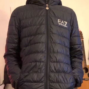 Svart pufferjacka från EA7 - Säljer en snygg mörkblå jacka från EA7. Jackan har en dragkedja framtill och är långärmad. Perfekt för kyligare dagar med sin quiltade design som håller värmen. Den har en stilren look med EA7-loggan på bröstet. Den har dock ett litet hål vid magen men inget man ser om man inte kollar noga.