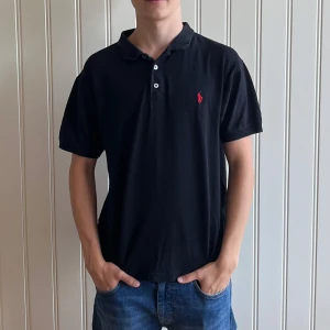 Svart pikétröja från Ralph Lauren - Säljer en klassisk svart pikétröja från Ralph Lauren med den ikoniska loggan broderad i rött på bröstet. Tröjan har korta ärmar och en stilren krage med knappar. Perfekt för en avslappnad men stilfull look. Storleken är M och skicket är mycket bra.