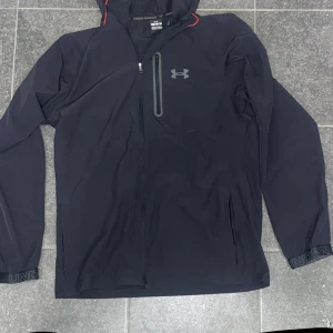 Svart jacka från Under Armour - Säljer en svart jacka från Under Armour med dragkedja och huva. Jackan har en diskret logga på bröstet och en ficka med dragkedja på bröstet. Perfekt för träning eller vardagsbruk. Behöver en ny dragkedja men annars helt ny