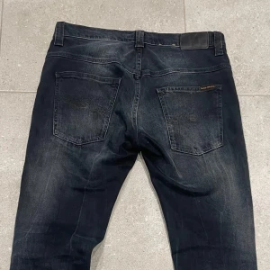 Svarta jeans från Nudie Jeans - Snygga svarta jeans från Nudie Jeans med klassisk femficksdesign och subtila slitningar. De har en knappgylf och en läderpatch baktill i midjan. Slim passform, 31W 34L. Bilder på, mått flera bilder kan fås vid intresse 