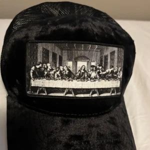 Svart keps med motiv av Nattvarden - Säljer denna keps från hatstore, med ett detaljerat motiv av Nattvarden på framsidan. Kepsen har ett nätmaterial på sidorna och ett stilrent märke på sidan. Perfekt för en unik stil.