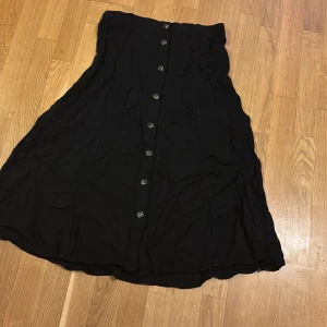 Svart kjol med knappar - Snygg svart kjol med knappar framtill. Kjolen har en elegant och flytande design som ger en fin siluett. Perfekt för att skapa en stilren look.