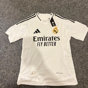 Vit fotbollströja från Adidas - Säljer en vit fotbollströja från Adidas med Real Madrid-logga och sponsortryck. Tröjan har korta ärmar och svarta detaljer på axlarna. Namnet 'Bellingham' och nummer 5 är tryckt på baksidan. Perfekt för fotbollsfans!