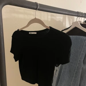 Svart kortärmad crop topp från Zara - Säljer en stilren svart topp från Zara. Den är kortärmad och har en klassisk design som passar till det mesta. Perfekt till jeans, crop toppen är i storlek S.