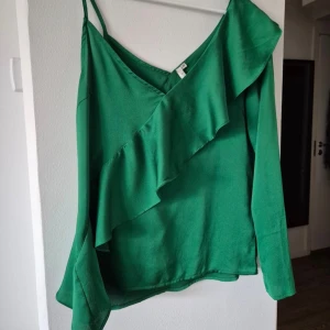 Grön blus med volangdetalj - Snygg grön blus med volangdetalj och smala axelband. Perfekt för en stilren look med en touch av elegans. Blusen har en asymmetrisk design och långa ärmar.