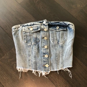 Jeans tubtopp med knappar - Snygg jeans tubtopp med knappar framtill och fransig nederkant. Perfekt för en cool och avslappnad stil. Den har en elastisk rygg för bättre passform.
