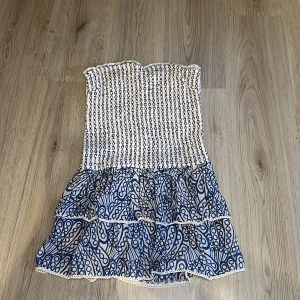 Vit och blå axelbandslös klänning från Gina Tricot - Säljer en söt axelbandslös klänning från Gina Tricot i vitt och blått. Klänningen har ett smockat liv och volangkjol med ett unikt mönster. Perfekt för sommarens varma dagar! 💙aldrig använt då den inte passar mig i smaken💙