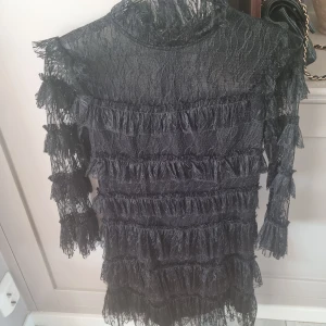 Svart spetsklänning med volanger - Snygg svart klänning i spets med volanger. Den har långa ärmar och en hög hals, vilket ger en elegant look. Perfekt för en kväll ute eller en speciell tillställning.