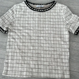 Vit och svart rutig topp från Zara - Snygg vit och svart rutig topp från Zara med korta ärmar. Den har pärldetaljer runt halsringningen och ärmsluten som ger en elegant touch. Perfekt för att lyfta vilken outfit som helst!