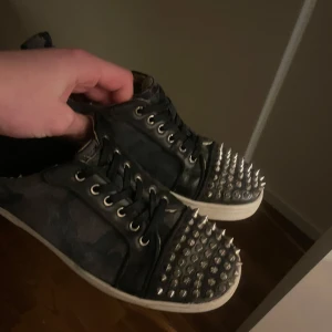 Camouflage loubs med nitar - Säljer ett par coola sneakers i camouflage med nitar på tån. Skorna har snörning och en unik design med röd sula. Perfekta för att ge din outfit en edgy touch.