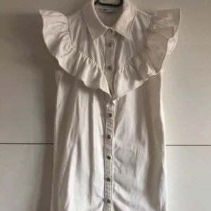 Vit klänning med volangdetaljer från Zara - Säljer en stilren vit klänning från Zara med volangdetaljer längs axlarna och knappar framtill. Perfekt för en chic och avslappnad look. Klänningen har en klassisk krage och är ärmlös.