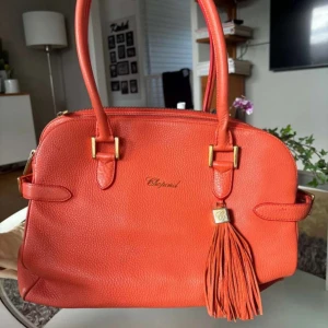 Korallfärgad Chopard handväska - Snygg orange handväska från Chopard i skinn med gulddetaljer och en dekorativ frans. Väskan har två handtag och en dragkedja upptill. Väskan är väldigt exklusiv och därför är varan väldigt dyr. Bilderna är väldigt suddiga men om du är intresserad kan jag ta nya bilder och skicka till dig