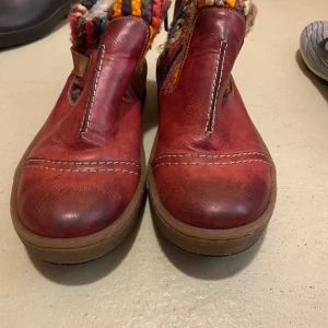 Röda boots från Rieker - Säljer ett par röda boots från Rieker med färgglada stickade detaljer runt ankeln. Skorna har en rund tå och en robust sula för extra komfort. Perfekta för att ge en färgklick till din outfit!