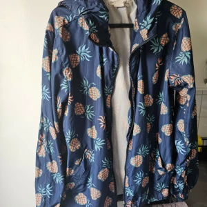 Mönstrad vindjacka från L.O.G.G. - Säljer en cool vindjacka från L.O.G.G. med ett unikt ananasmönster i orange och grönt på en mörkblå bakgrund. Jackan har dragkedja och huva, perfekt för en stilren look. Passar bra för vår och sommar.