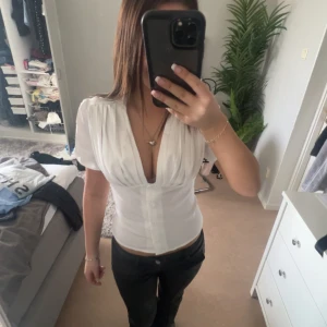 Vit blus med v-ringning - Säljer en stilren vit blus med v-ringning och korta puffärmar. Blusen har en figurnära passform och knappar framtill. Perfekt för både vardag och festliga tillfällen.