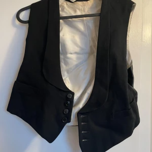 Svart väst med knappar - Snygg svart väst med knappar framtill och en elegant krage. Perfekt för att ge en stilren touch till din outfit. Passar bra till både formella och avslappnade tillfällen.
