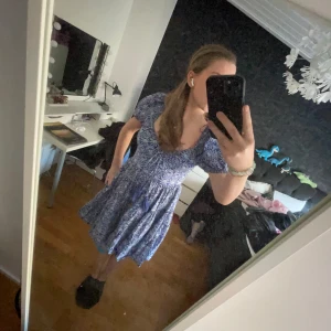 Blå blommig klänning - Charmig blå klänning med blommigt mönster och korta ärmar. Klänningen har en smickrande passform med volangdetaljer och en v-ringad hals. Perfekt för sommarens alla tillfällen.