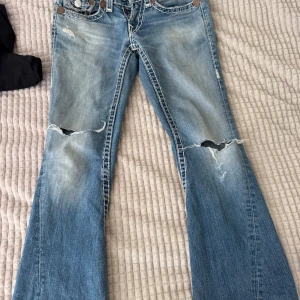 Blå bootcut jeans från True Religion - Snygga blå bootcut jeans från True Religion med slitna detaljer och hål på knäna. Jeansen har en klassisk femficksdesign och kontrastsömmar. Perfekta för en avslappnad stil med en touch av edge.