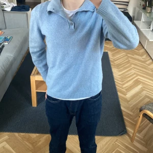 Ljusblå tröja från Massimo Dutti - Säljer en stilren ljusblå tröja från Massimo Dutti i en mjuk ull- och kashmirblandning. Tröjan har en ribbad krage med knappar och långa ärmar. Perfekt för kyligare dagar!