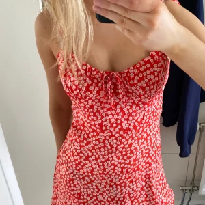 Röd blommig klänning med knytband - Charmig röd klänning med vita blommor och knytband vid axlarna. Den har en smockad överdel och volangdetaljer vid fållen. Perfekt för sommarens varma dagar.