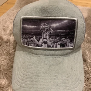 Grå Messi velvet keps  - Snygg grå keps från Berretto med ett unikt fototryck med messi på framsidan. Kepsen har en klassisk design med nätpaneler på sidorna för extra ventilation. Perfekt för en avslappnad stil.