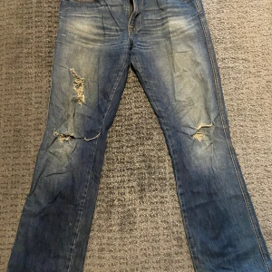 Blå jeans från Nudie Jeans - Säljer ett par blå jeans från Nudie Jeans med slitna detaljer 