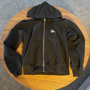 Svart huvtröja från Stüssy - Säljer en svart huvtröja från Stüssy med dragkedja och stor logga på ryggen. Tröjan har långa ärmar och en praktisk ficka framtill. Perfekt för en avslappnad stil.