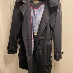 Svart trenchcoat från Esprit  - Snygg svart trenchcoat från Esprit med klassisk design. Jackan har ett bälte i midjan och knappar framtill. Den är långärmad med justerbara spännen vid ärmsluten. Insidan har ett randigt foder i blått och vitt. Perfekt för höst och vår!