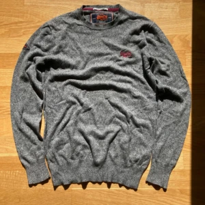 Grå tröja från Superdry - Säljer en grå tröja från Superdry i bomull.. Tröjan har en broderad logga på bröstet och en klassisk rund halsringning. 