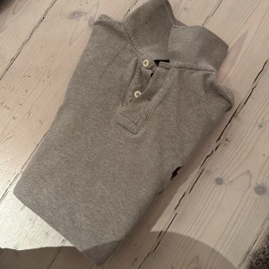 Grå pikétröja från Ralph Lauren - Säljer en stilren grå pikétröja från Ralph Lauren i slim fit. Tröjan har korta ärmar och en klassisk krage med knappar. Perfekt för en avslappnad men ändå snygg look. 🐎