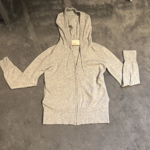 Grå kashmir hoodie från Absolut Cashmere - Säljer en supermjuk och stilren grå hoodie i kashmir från Absolut Cashmere. Den har en dragkedja framtill och långa ärmar, perfekt för kyliga dagar. Huvan ger en avslappnad look och den ribbade nederkanten och ärmsluten ger en snygg finish. Den har ett hål vid vänster arm som inte så stort men inget annat är sönder. Perfekt för både mys och stil! 🔥