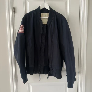 Svart bomberjacka från Ralph Lauren - Säljer en svart bomberjacka från Ralph Lauren med en amerikansk flagga på ärmen. Jackan har dragkedja och ribbade muddar vid ärmslut och nederkant. Perfekt för en stilren look.