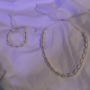 Halsband och armband med snäckor - Snyggt set med halsband och armband dekorerade med vita snäckor och små guldfärgade pärlor. Perfekt för en strandlook eller sommarfest. Justerbara band för perfekt passform.