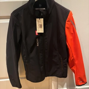 Svart och orange racingjacka från Aprilia - Snygg racingjacka från Aprilia i svart med en kontrasterande orange ärm. Jackan har dragkedja framtill och två praktiska fickor med dragkedjor. Perfekt för racingentusiaster som vill ha stil och funktionalitet.