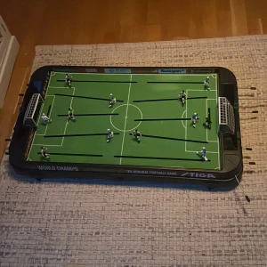 Fotbollsspel från Stiga - Säljer ett klassiskt fotbollsspel från Stiga. Spelplanen är grön med vita linjer och har två lag med spelare i blått och vitt. Perfekt för fotbollsälskare som vill ha kul inomhus!