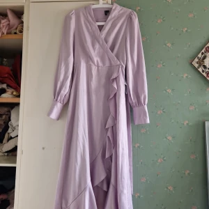 Ljuslila omlottklänning - Säljer en elegant ljuslila omlottklänning med långa ärmar och volangdetaljer framtill. Klänningen har en v-ringad design och ett knytband i midjan för en smickrande passform. Perfekt för vårens och sommarens tillställningar!