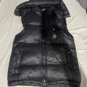 Svart dunväst från Moncler - Snygg svart dunväst från Moncler med dragkedja och tryckknappar. Västen har en hög krage och en logotyp på bröstet. Perfekt för kyliga dagar när du vill hålla stilen. 🖤