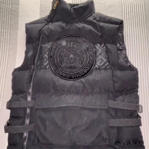 Svart dunväst - Snygg svart dunväst från Glorious Gangsta med en stor broderad logga på bröstet. Västen har en dragkedja och justerbara remmar i sidan för perfekt passform. Insidan har ett mönster som ger en extra touch av stil.