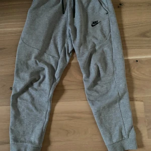 Grå mjukisbyxor från Nike - Säljer ett par grå mjukisbyxor från Nike med elastisk midja och ribbade muddar. Byxorna har sidofickor och en Nike-logga på benet. Perfekta för en avslappnad stil.