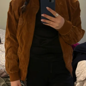 Brun mockajacka från Petroff - Säljer en snygg brun vintage mockajacka från 80- talet och märke Petroff med dragkedja och ribbade muddar. Jackan har en cool vintage look och är perfekt för höst och vår. Insidan har ett randigt mönster som ger en extra touch av stil.