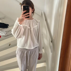 Vit blus med volangdetaljer - Säljer en söt vit blus med volangdetaljer runt halsen. Blusen har långa ärmar med elastiska muddar och en lös passform. Perfekt för en stilren och bekväm look.