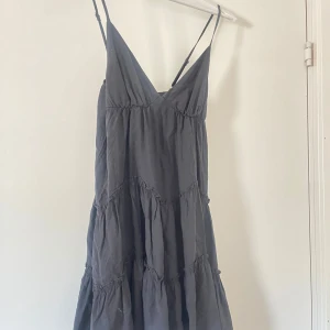 Svart axelbandslös klänning - Säljer en stilren svart klänning med smala axelband och v-ringning. Klänningen har en luftig passform med volangdetaljer som ger en fin rörelse. Perfekt för varma dagar eller sommarens fester.