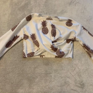 Vit långärmad topp med ananasmönster - Säljer en vit långärmad topp med brunt ananasmönster. Toppen är croppad och har en unik twistdetalj framtill. Perfekt för en somrig look!