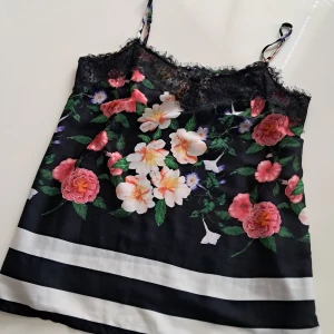 Blommig linnetopp från Hallhuber - Snygg blommig linnetopp från Hallhuber i svart med färgglada blommor och spetsdetaljer vid halsen. Toppen har smala axelband och ett randigt mönster längst ner. Perfekt för en somrig look!