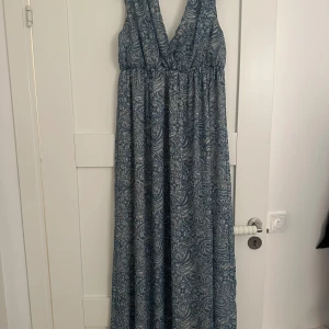 Blå mönstrad långklänning - Säljer en vacker blå långklänning med ett detaljerat paisleymönster. Klänningen har en v-ringad design och är ärmlös, vilket ger en elegant och luftig känsla. Perfekt för sommarens varma dagar.